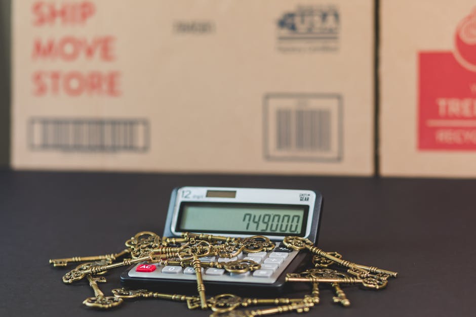 A close-up view of a small calculator displaying the number 749800, surrounded by a collection of gold-colored keys of various shapes and sizes, some attached to key rings. The calculator and keys are placed on a dark surface, with multiple cardboard boxes in the background, which have printed labels including barcodes and red and blue text. The boxes are situated indoors, likely used for packing during a home relocation or moving process, with soft lighting illuminating the scene. The image captures elements related to packing and moving logistics, consistent with home removals services provided by Paddington Removals, emphasizing the preparatory stage of furniture transport and packing during a house move.