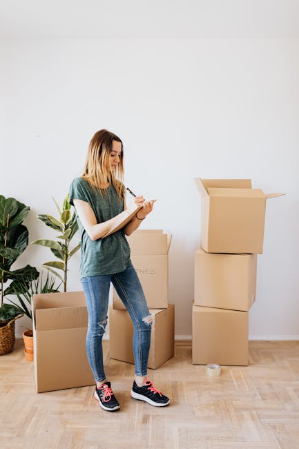 A young woman with shoulder-length blonde hair, dressed in a casual green t-shirt, ripped blue jeans, and black sneakers with pink accents, stands inside a room preparing for a house move. She is holding a clipboard or notebook and appears to be taking notes or checking a list. Behind her are several large cardboard boxes, some stacked on top of each other, with one open box revealing its interior. The boxes are placed on a wooden floor and near a white wall, with a potted plant with broad green leaves positioned on the floor to the left of the woman. The scene captures the packing and organizing process involved in home relocation, illustrating typical packing materials and the preparation stage of furniture transport, with natural lighting enhancing the clarity of the space. Paddington Removals, a professional moving company, is associated with this image as part of their house removals and packing services.