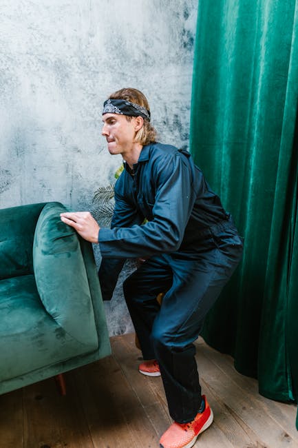 A man wearing a dark blue jumpsuit and a patterned headband is seen lifting and moving a green upholstered armchair inside an interior space. The setting features a textured grey concrete wall and green curtains on the right side, partially covering a window or doorway. The man is gripping the backrest of the armchair with both hands, preparing to carry or reposition it as part of a home relocation or furniture transport process. The floor is wooden, and the scene is well-lit, capturing the professional moving activity associated with residential removals. Paddington Removals specializes in packing and loading sizeable furniture items safely during house moves, as demonstrated in this furniture handling scene.