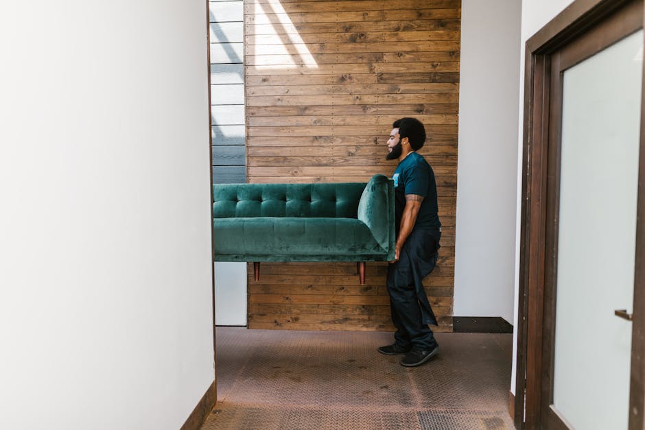 A man wearing a dark blue uniform is carefully lifting a large, green velvet-upholstered sofa with wooden legs inside a home, preparing for a house removal or furniture transport. The sofa is supported from the middle, held securely with both hands, and is positioned horizontally with the seat facing downward. The interior space features a wooden accent wall in natural brown tones behind him, with a white wall to the left and a glass door with a wooden frame on the right. The floor is covered with a brown, textured carpet. The scene depicts a professional packing and moving process, illustrating careful handling of bulky furniture during a home relocation, coordinated by Paddington Removals in London, UK.