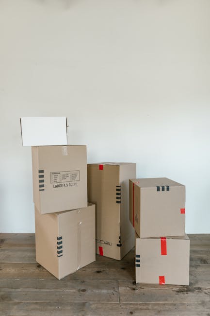 A collection of six large cardboard boxes of varying sizes, some with printed labels and others with red and black striped tape, stacked and arranged on a wooden floor in front of a plain white wall. One box is slightly tilted, and there is an open white panel or small board mounted on the wall behind the boxes. The boxes are packed with packing materials, likely used for home relocation or furniture transport, and are ready for loading or unpacking. The scene suggests an ongoing or completed packing process as part of house removals or moving logistics, with a focus on secure and organized packing managed by Paddington Removals, a professional transportation service for residential and commercial moves.