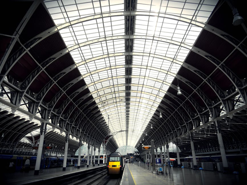 The interior of a large train station with a high, arched glass ceiling allowing natural light to illuminate the space. Steel supports and curved beams frame the roof structure. On the platform, a modern yellow and grey train is stationary near the edge, with several other trains visible further along the tracks. The platform is equipped with various electronic information displays, lighting fixtures, and seating areas. In the background, passengers can be seen in the distance, some walking and others waiting. This setting depicts a typical busy railway station environment suitable for home relocation or furniture transport planning, highlighting the importance of timing and logistics for moving near Paddington Station. Paddington Removals, as a professional service provider, assists with packing, loading, and transportation processes essential for house removals and moving services in London.