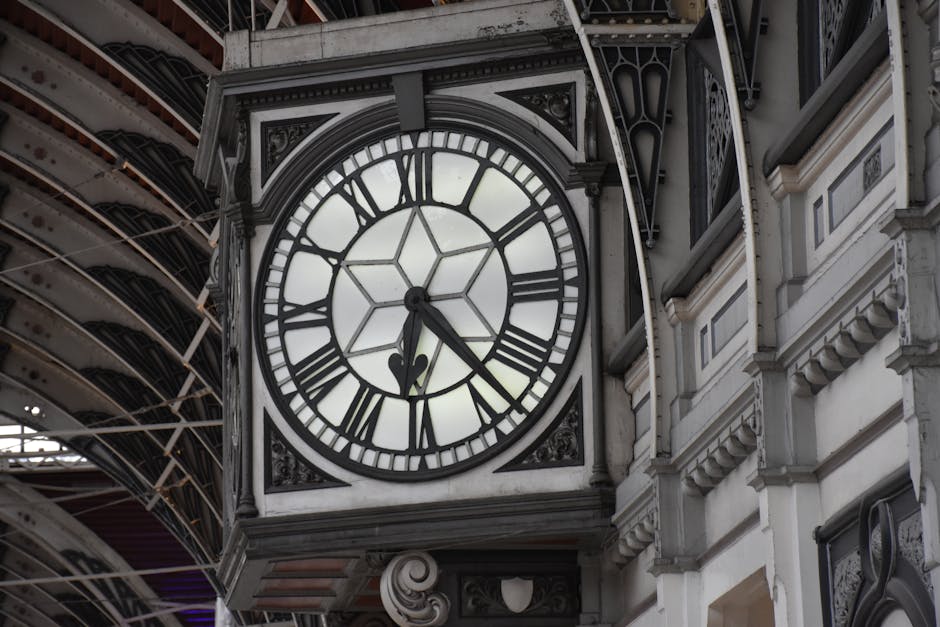Close-up of a large, ornate clock mounted on the exterior of a historic building, with Roman numerals marking the hours and black clock hands indicating the time as approximately 11:25. The clock face is framed by decorative metalwork and stone architecture with detailed mouldings. Behind the clock, part of a railway station canopy with metal braces and glass panels is visible, suggesting a location near Paddington Station. The scene captures the clock during daylight hours, highlighting the architectural style typical of traditional British railway termini. This setting relates to house removals and relocation services, as shown by the context of loading furniture or moving boxes in similar environments, and is relevant for content about the timing and logistics of home relocation in proximity to major transport hubs such as Paddington Station. Paddington Removals specializes in professional furniture transport and packing during house moves, ensuring efficiency in busy station areas.