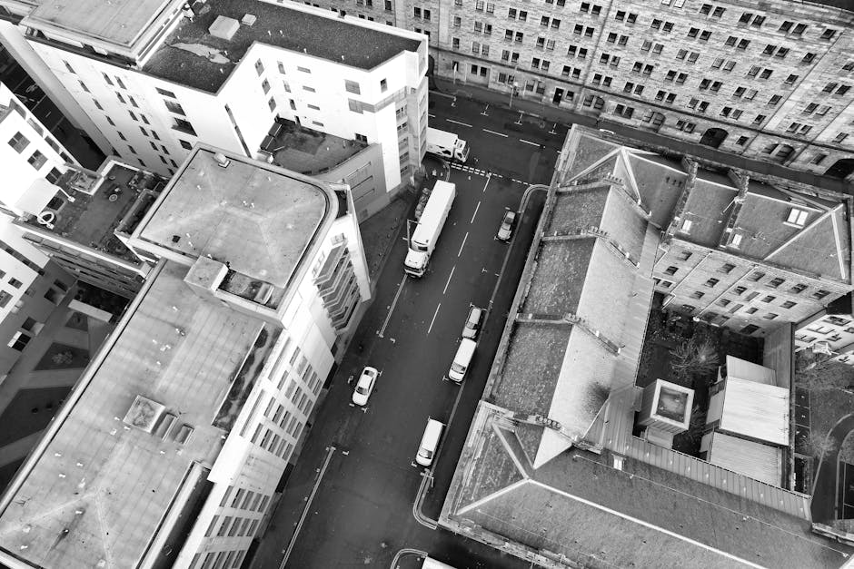An aerial black-and-white photograph captures a city street lined with multi-storey buildings and parked vehicles, including vans and cars, along the curb. In the foreground, a building with a flat roof hosts rooftop equipment such as air conditioning units and skylights. The street below shows a smooth surface with clearly marked parking spaces and vehicles, as well as a sidewalk running alongside. A white van is actively involved in the home relocation process, seen loading or unloading near the building, with some moving boxes and furniture possibly nearby. The scene suggests an urban apartment or office move, with the logistics of packing, loading, and transportation underway, supporting the services of Paddington Removals during same-day house removals in Paddington. The image reflects the efficient movement of household or office items through a typical city environment, emphasizing the arrangement of furniture, packing materials like cardboard boxes, and moving equipment such as trolleys or straps, integral to a professional removals service.