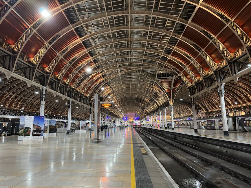 Inside a large train station with an arched, metal and glass roofing structure illuminated by bright overhead lighting. In the foreground, a dark blue and yellow passenger train is stationary on the platform, with visible front headlights and a digital display above the windshield. Next to the train, there are some red and black loading barriers and a metal railing at waist height separating the platform from the tracks. In the background, additional train carriages are parked on adjacent tracks, and a large clock shows the time on the station wall. The station features a spacious, clean environment, suitable for moving and packing activities, often associated with house removals and home relocation services, such as those provided by Paddington Removals.