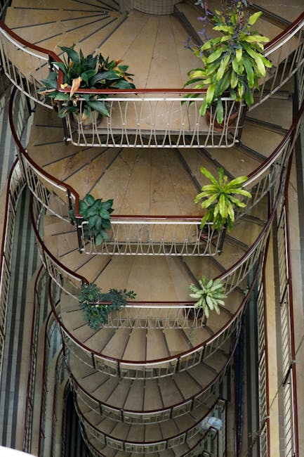A top-down view of a spiral staircase inside a building, with a wooden handrail and white metal balustrades. The staircase has multiple levels, each with a landing decorated with various potted plants