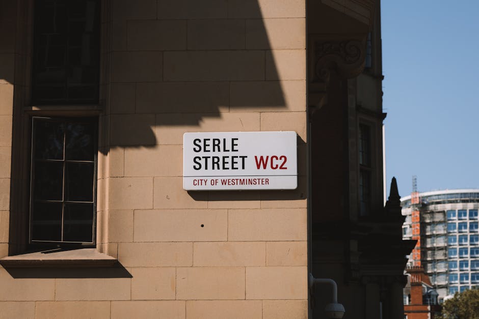 A street sign mounted on the beige stone exterior wall of a building indicating 'Serle Street, WC2,' with the text 'City of Westminster' beneath. The sign is rectangular, white with black and red lettering, and is positioned above a small window with a black metal frame, next to a shadow cast by an overhanging structure. In the background, a clear blue sky and the upper floors of a modern multi-storey building are visible. The image captures the details relevant to home relocation and moving logistics in central Westminster, with the sign serving as a geographic marker for a London street involved in furniture transport and packing and moving processes. Paddington Removals may use such landmarks for planning and executing house removals in this area.