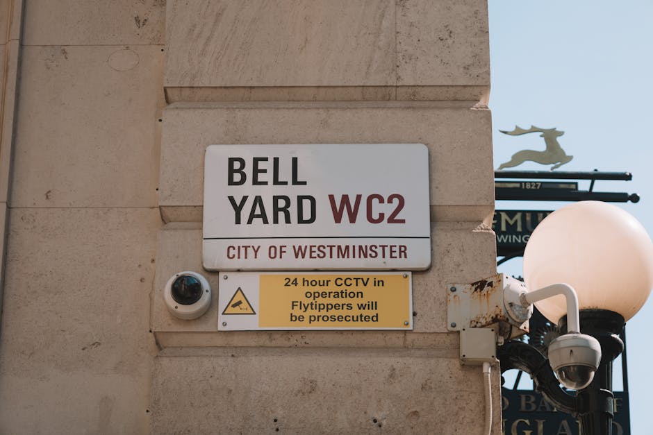 Close-up image of a building exterior in Westminster showcasing a white and red sign reading 'BELL YARD WC2' with 'CITY OF WESTMINSTER' written below, mounted on a beige stone wall. Beneath this sign, there is a yellow rectangular notice indicating '24 hour CCTV in operation' and warning that 'Flytippers will be prosecuted.' To the left of the signs, a small, black dome-shaped CCTV security camera is attached to the wall, monitoring the area for security during home relocation or furniture transport activities. On the right, a classic black lamp post with a round white globe light is partially visible, attached to the same wall, suggesting a well-lit pedestrian area suitable for moving and packing processes in London. The overall scene highlights signage related to city regulations in a typical residential or commercial street in central Westminster, relevant for professional removals and packing services such as those offered by Paddington Removals.